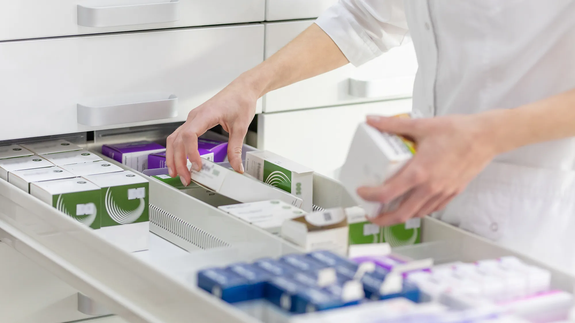 Die Hände eines Apothekers ordnen verschiedene Medikamentenschachteln in einem gut beleuchteten Apothekerschrank. Die Szene vermittelt Präzision und Sorgfalt in einer medizinischen Einrichtung.