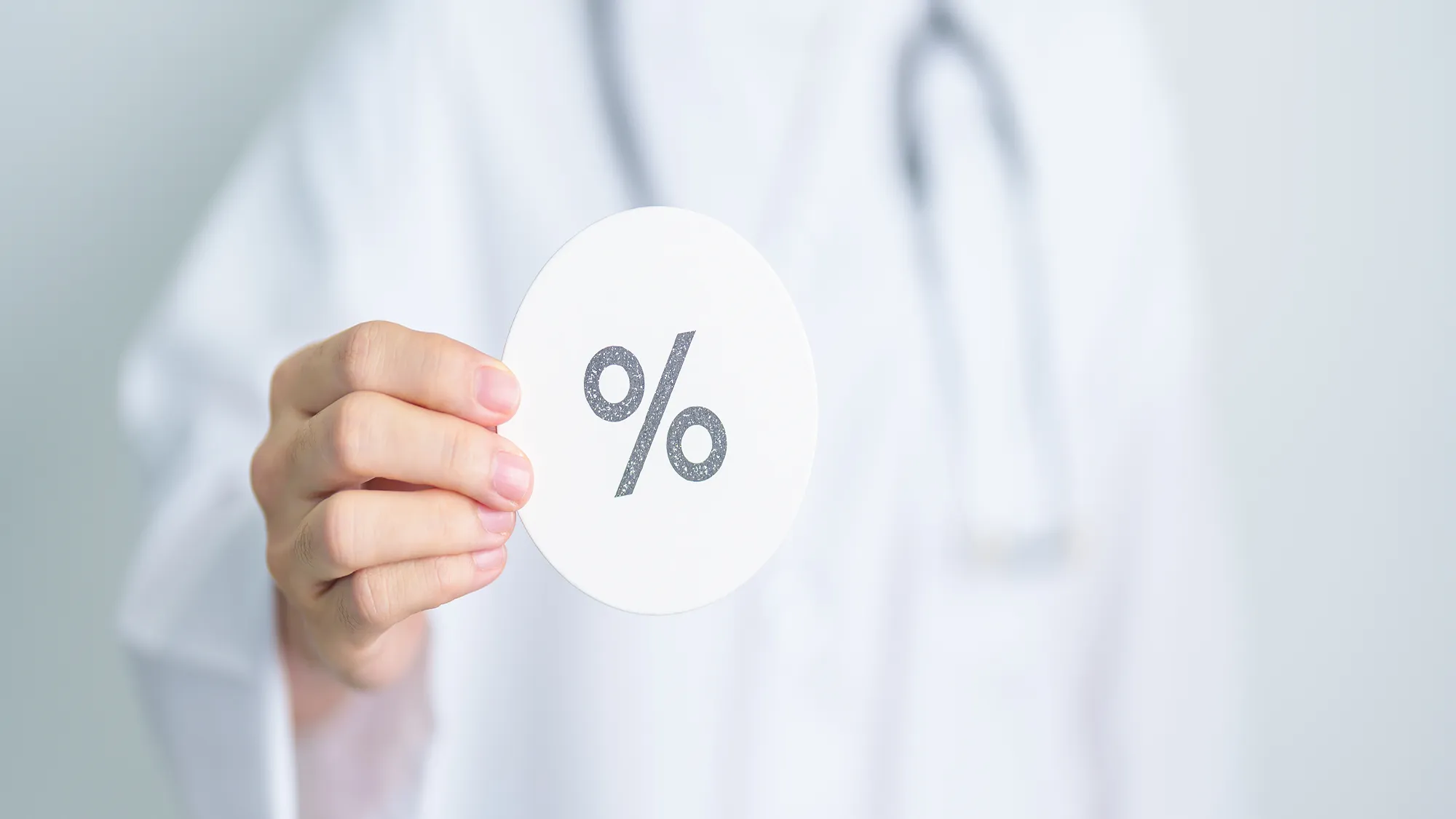 Ein Apotheker oder eine Person in einem weißen Kittel hält eine runde Karte mit einem Prozentzeichen in der Hand. Der Hintergrund ist weich und unscharf, was auf eine professionelle oder medizinische Umgebung hindeutet.
