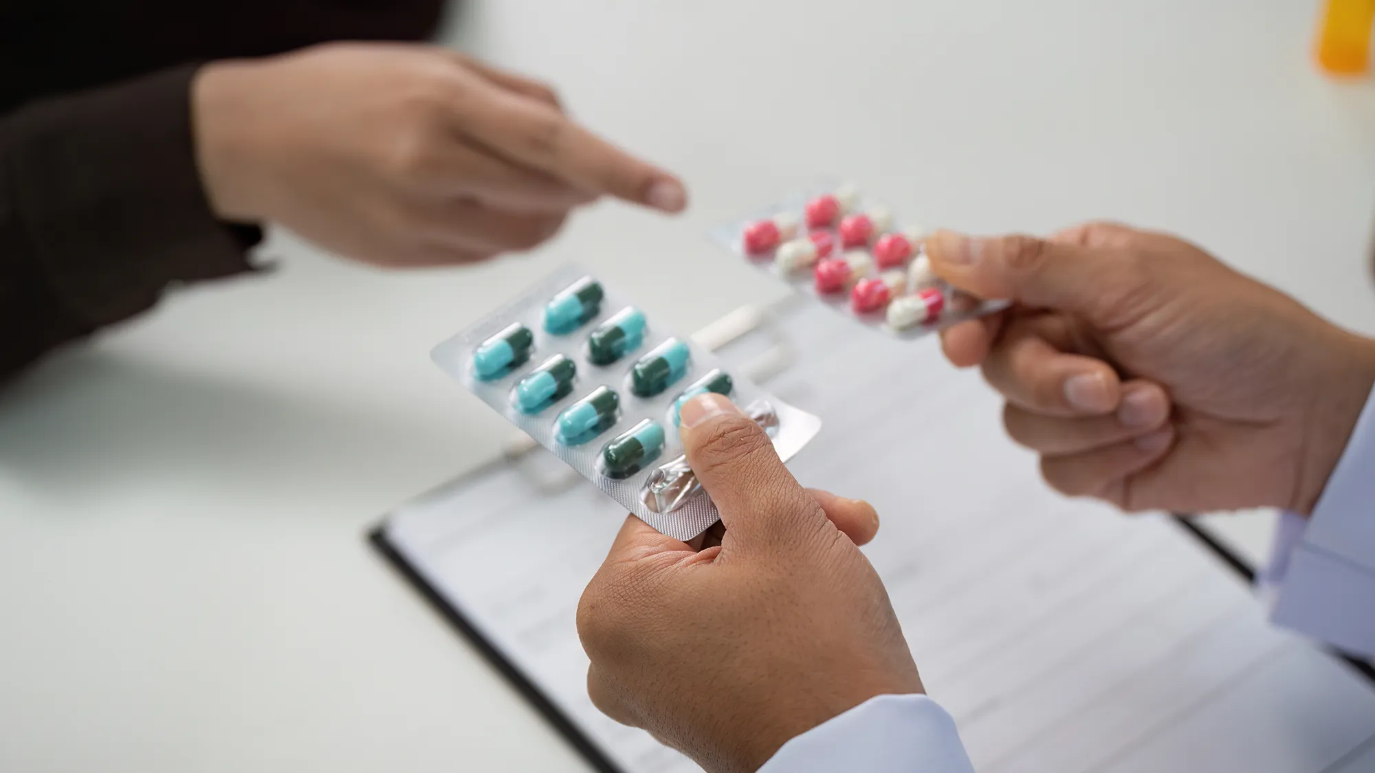 Close-up of two hands exchanging blister packs of pills, one with green capsules and the other with pink pills, suggesting a medical consultation.