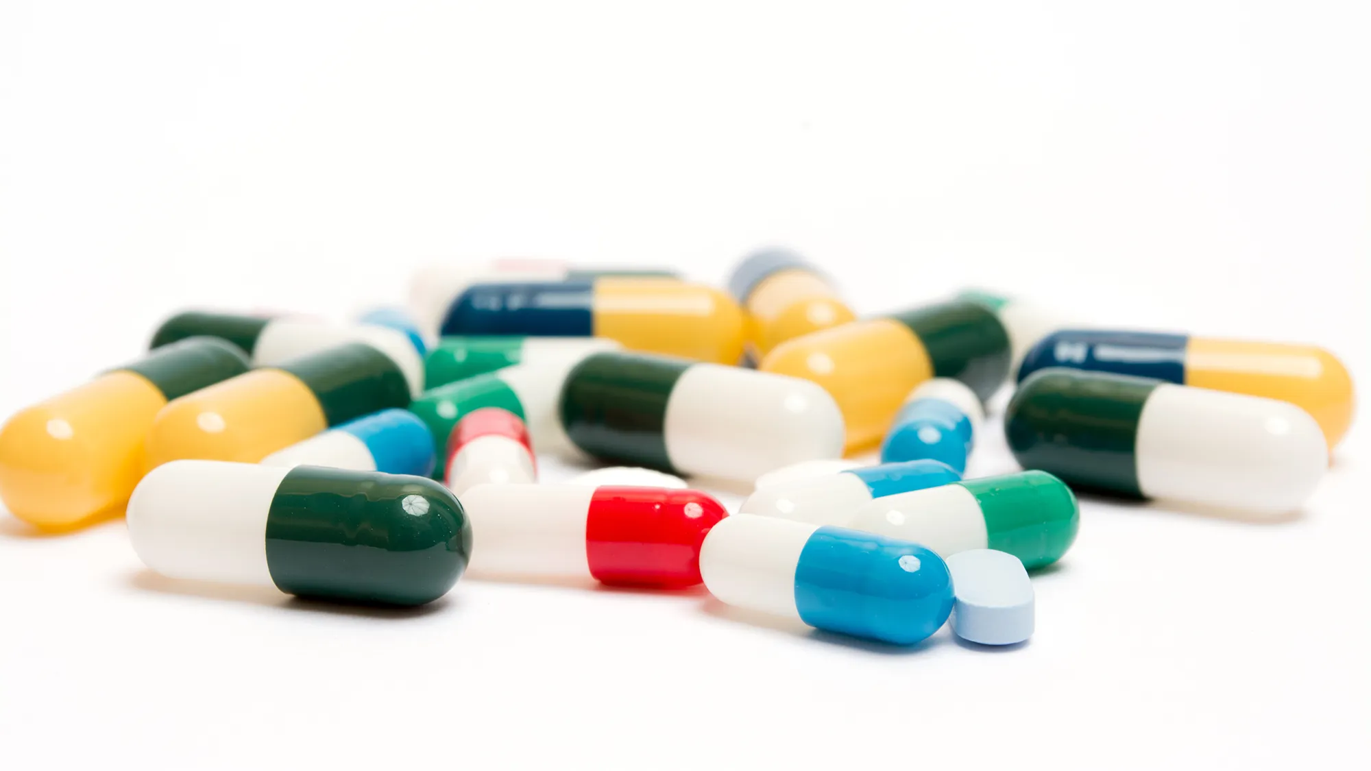 Ein Haufen bunter Kapseln und Tabletten, verstreut auf einer weißen Fläche. Die Pillen haben verschiedene Farben, darunter grün, blau, rot und gelb.